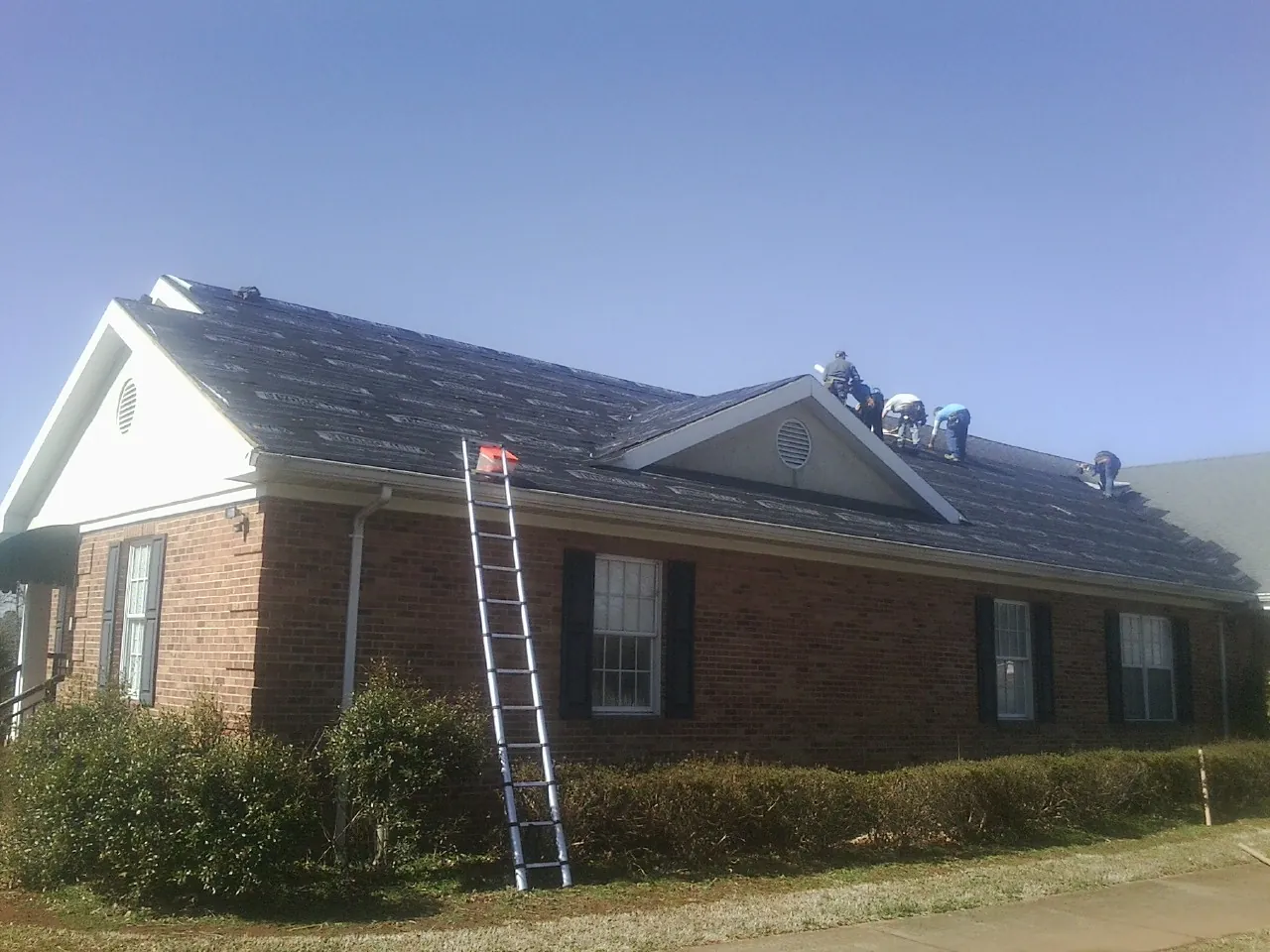 Crew tearing shingles off a brick ranch roof with a ladder set — Emergency Roof Repair work by our crew in Skippers