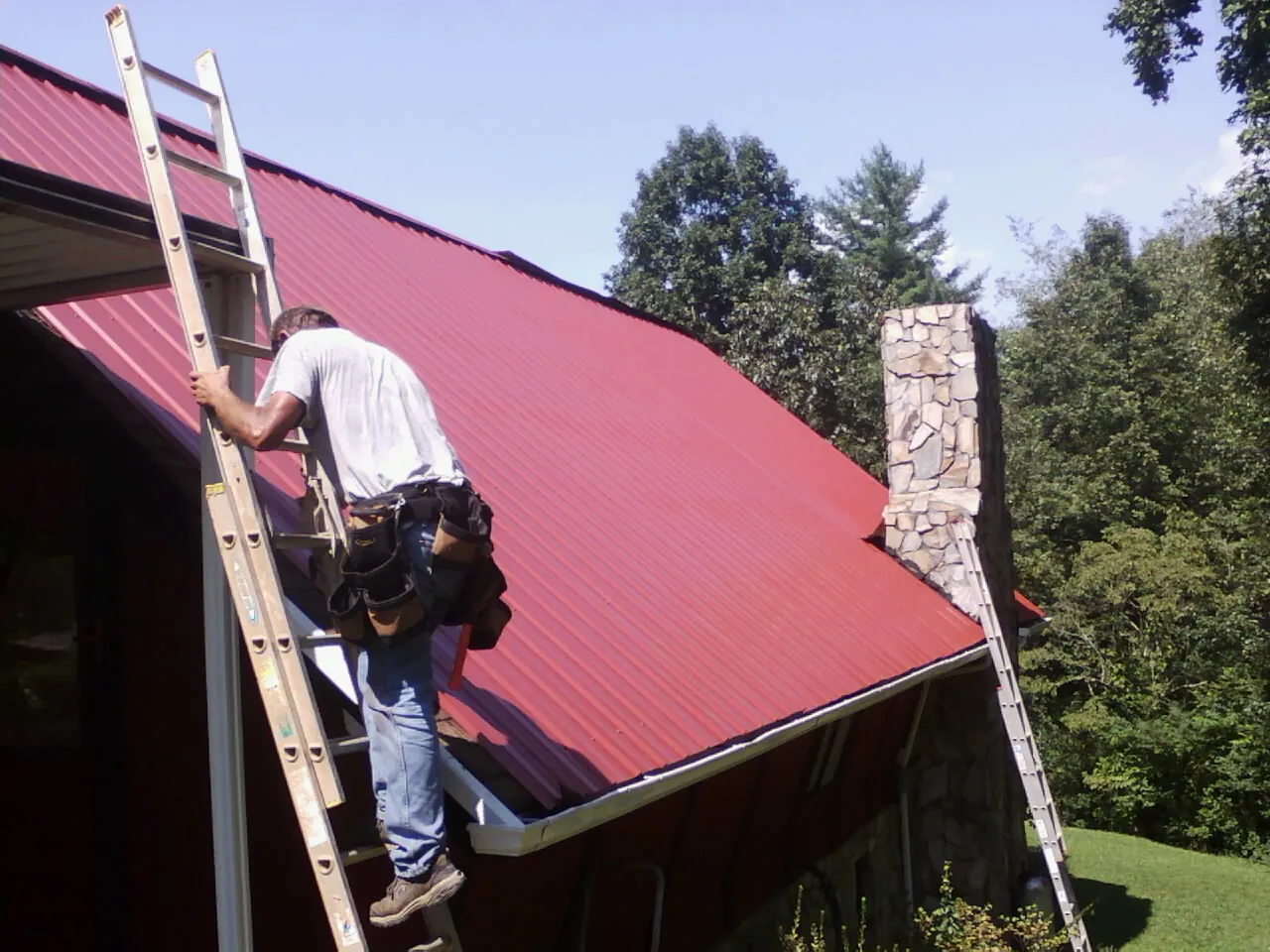 Roofer on a ladder beside a red metal roof and stone chimney — Soffit & Fascia Repair by Roofing Brothers in Skippers